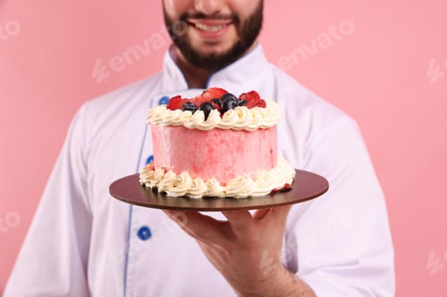 Preview: Happy confectioner in uniform holding cake with berries on pink background, closeup