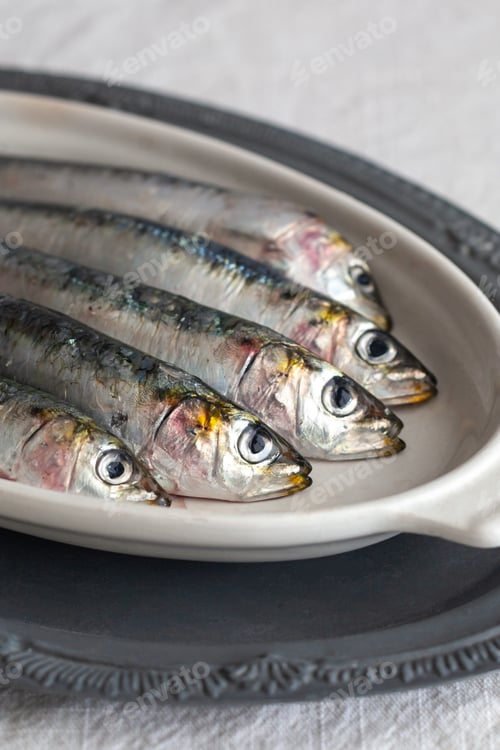 Preview: A close up picture of fresh, raw sardines on a white plate, vertical