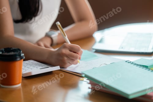 Preview: Businesswoman writing notes while working at desk with laptop and calculator