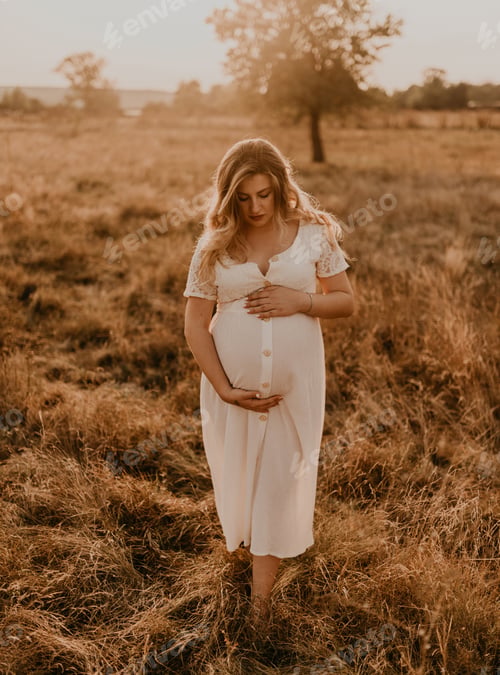 Preview: Caucasian pregnant young blonde woman in cotton white linen dress stand walking in meadow
