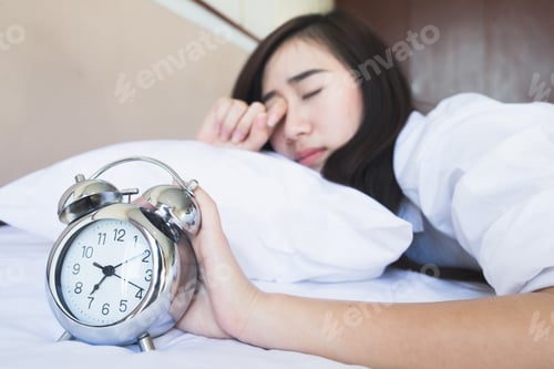 Preview: woman in bed trying to wake up with alarm clock.