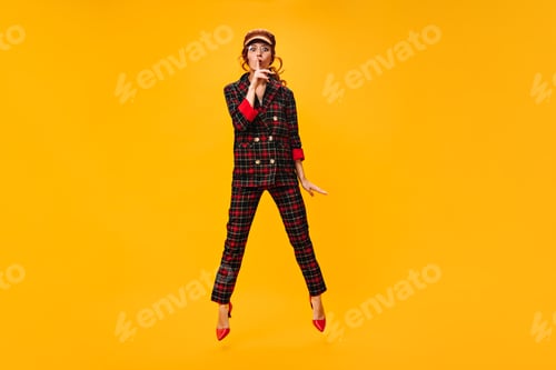 Preview: Attractive woman jumps on orange background and asks to keep secret. Fashionable girl in brown tren