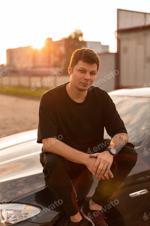 Preview: Fashionable young man model in stylish black summer clothes sits on the car at sunset