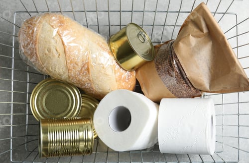 Preview: Shopping basket with products and toilet paper rolls on grey table, top view. Panic caused by virus