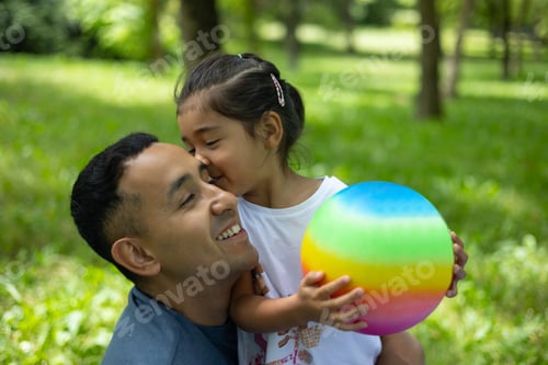 Preview: Little girl kissing her dad on the cheek while holding a rainbow colored ball in a green park