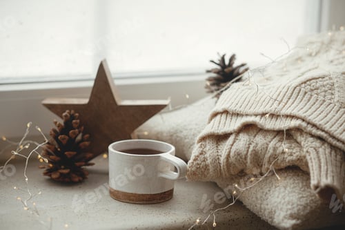 Preview: Stylish cup of tea with cozy knitted sweater, pine cone, wooden star and golden lights on windowsill