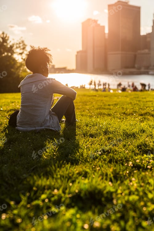 Preview: USA, New York City, Brooklyn, woman sitting on meadow looking at the skyline