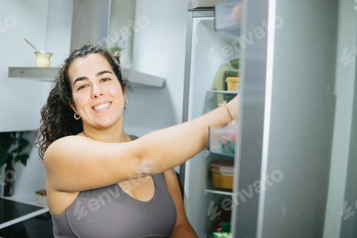 Preview: Curly woman doubting about what to eat from the fridge at the kitchen home to lose weight