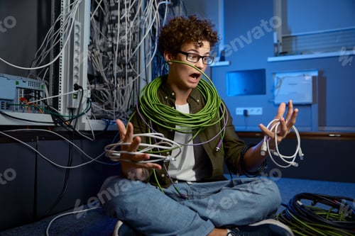 Preview: Shocked teenager geek wrapped in wire sitting on floor in data center