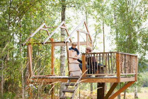 Preview: Two children playing on a wooden treehouse amidst lush greenery.