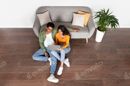 Preview: Indian couple using laptop sitting on floor, browsing web, above view