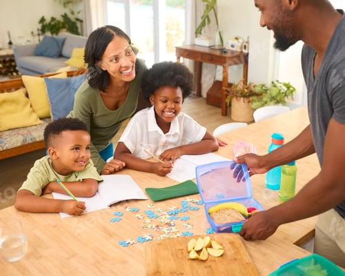 Preview: Mother Helping Children With Homework Sitting At Table As Father Makes Packed Lunch