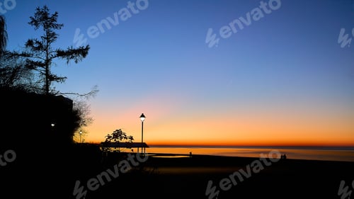 Preview: Beautiful colorful sunset sky over a tranquil body of water with silhouettes of street lamps