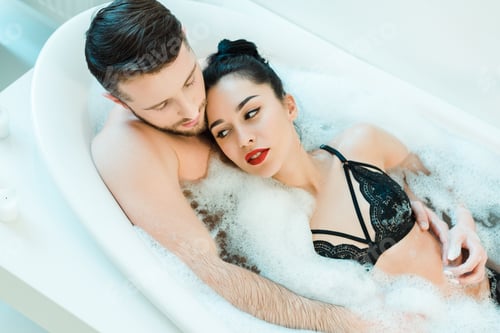 Preview: overhead view of handsome man lying in bathtub with attractive brunette woman in lace bra