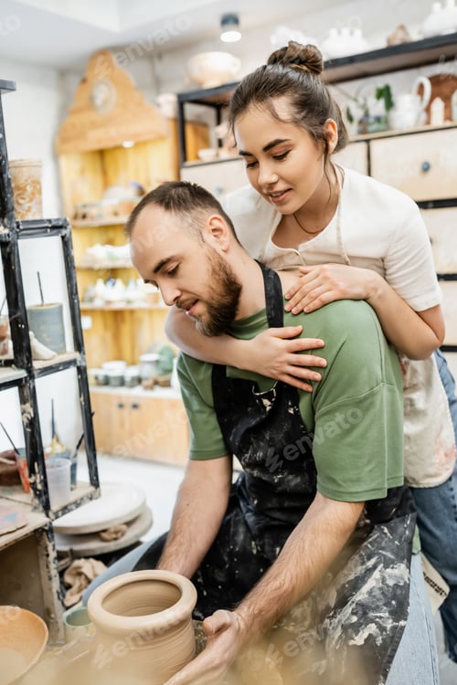 Preview: Smiling female potter in apron hugging boyfriend working with clay and pottery wheel in workshop