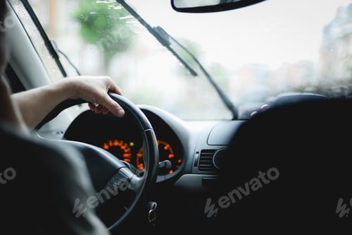 Preview: A young caucasian guy keeps his hand on the steering wheel while driving a car on a rainy day
