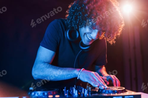 Preview: Man with curly hair using DJ equipment and standing in the dark neon lighted room