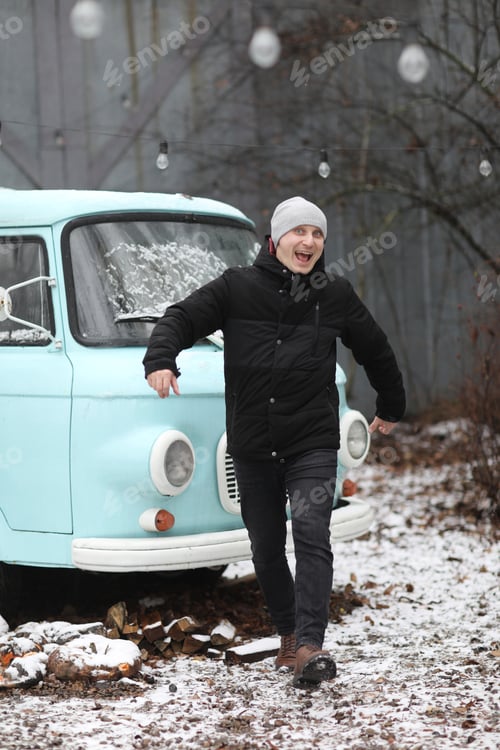 Preview: cheerful young man in winter near the retro blue car and light bulbs.
