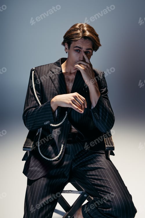 Preview: stylish man in trendy suit with pearl chain sitting on camp chair while looking at camera on dark