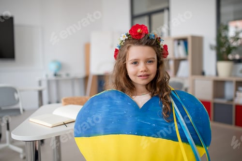 Preview: Little Ukrainian schoolgirl holding heart in Ukrainian colors and looking at camera at school