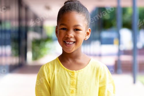 Preview: Portrait of smiling biracial schoolgirl standing in sunny school corridor with copy space
