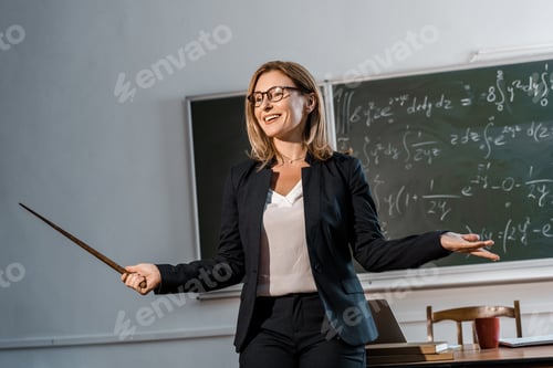 Preview: smiling female teacher with wooden pointer explaining mathematical equations in classroom