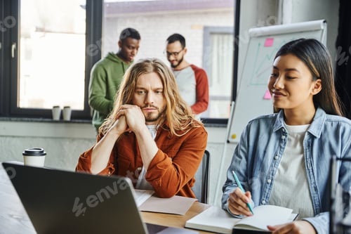 Preview: thoughtful long haired businessman looking at laptop near smiling asian woman with notebook while