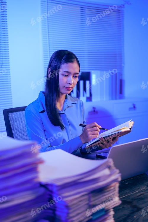 Preview: An Asian woman works late at night in the office, busy stressed while checking documents her desk