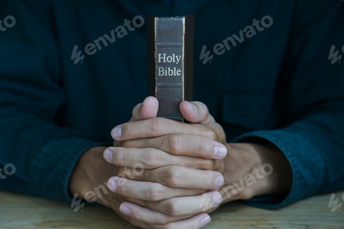 Preview: Christian man holding bible. Hands folded in prayer on a Holy Bible on wooden table.
