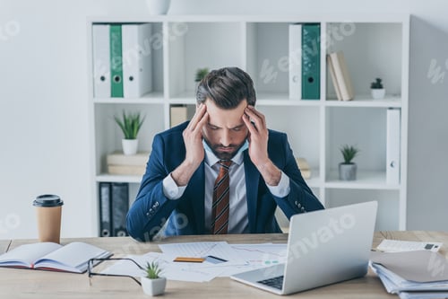 Preview: depressed businessman touching bowed head while sitting with closed eyes at workplace