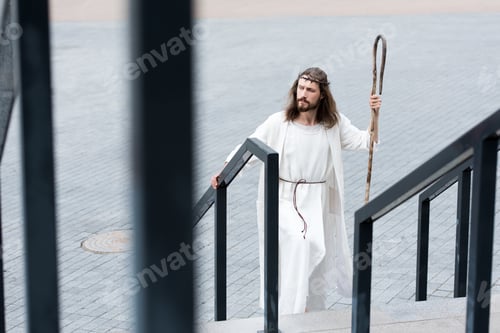 Preview: high angle view of Jesus in robe and crown of thorns walking on stairs with staff