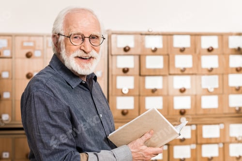 Preview: smiling senior male archivist holding book and looking at camera