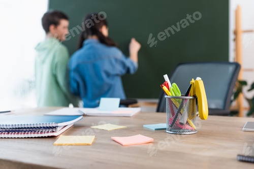 Preview: selective focus of school desk with notebooks and sticky notes near blurred classmates writing on