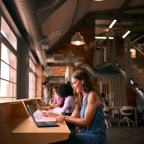 Preview: Businesswoman Working In Modern Multi-Cultural Office Listening To Music On Earphones Or Earbuds