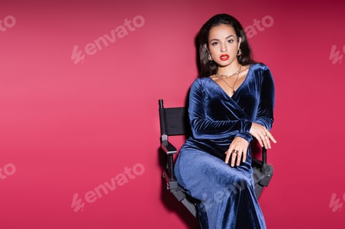 Preview: young brunette woman in blue velvet dress looking at camera while sitting on red background