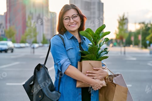 Preview: Middle aged woman with paper shopping bags with buying plant, looking at camera