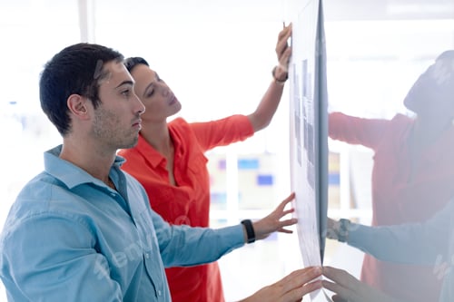 Preview: Side view of Caucasian architects discussing over blueprint on glass board in a modern office
