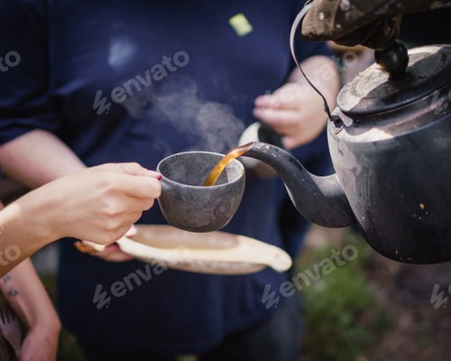 Preview: Closeup of a person pouring hot tea from a pot into a ceramic cup under the sunlight