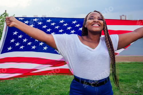 Preview: Smiling afro american woman holding USA flag and looking at camera autumn evening