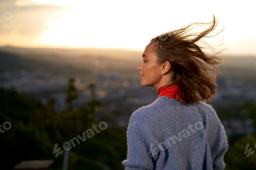 Preview: Woman enjoying a serene sunset over a cityscape with her hair blowing in the wind.