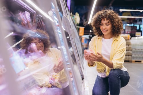 Preview: Young Woman Choosing Food Products in Refrigerated Section of Supermarket