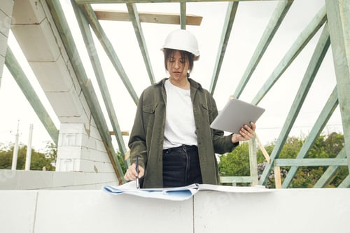 Preview: Stylish woman architect or engineer with tablet checking blueprints at wooden roof framing of house