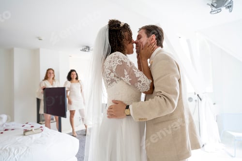 Preview: Bride and groom embracing and kissing in a bright room with bridesmaids in the background.