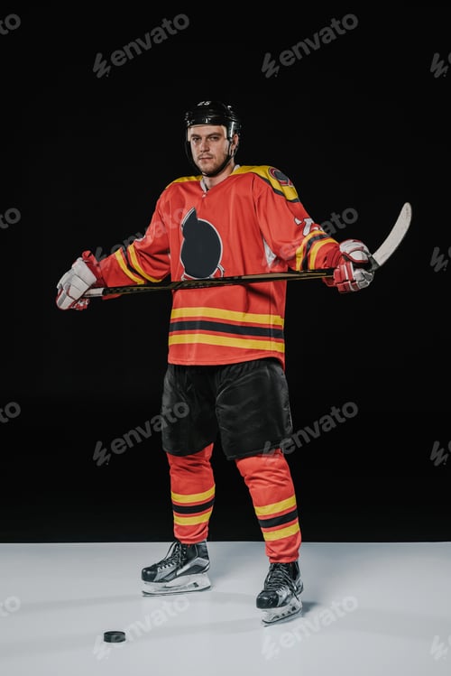Preview: full length view of professional ice hockey player holding hockey stick and looking at camera on
