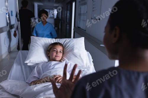 Preview: Diverse female doctor talking with girl lying in bed in hospital corridor