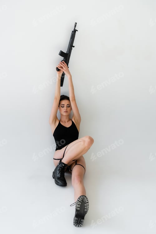 Preview: sensual woman holding rifle in raised hands while sitting with closed eyes on grey background