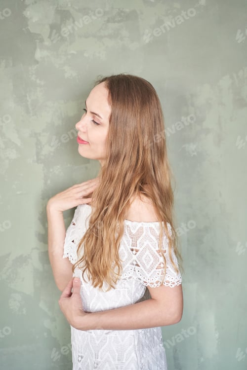 Preview: Young caucasian female in white lace dress posing against textured green wall