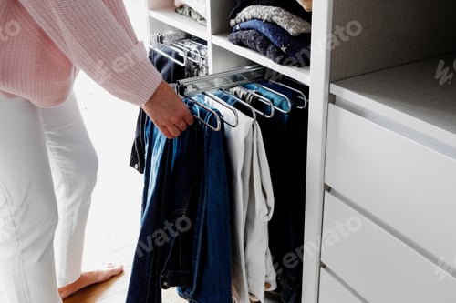 Preview: Woman hanging trousers in closet, organizing and ordering in wardrobe, neutral, pastel tones, jeans