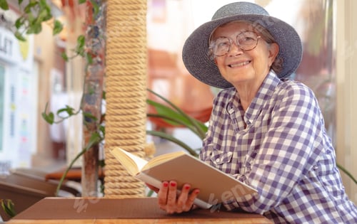 Preview: Portrait of beautiful senior woman with hat sitting at cafe table reading a book
