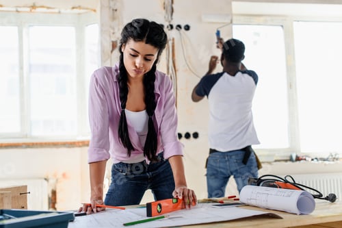 Preview: pensive young woman with spirit level looking at blueprint while her boyfriend working behind during